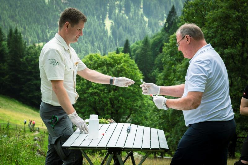 Zwei Forscher stehen an einem Tisch und hantieren mit Pipette und einem Behälter. Im Hintergrund bewaldete Hügel