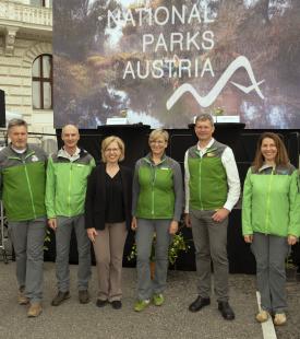 Eine Gruppe von acht Personen steht vor einer kleinen Bühe unter dem Slogan 'Nationalparks Austria'