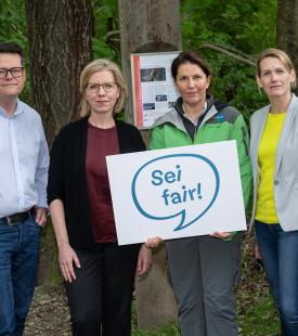 Drei Personen sind vor den Stämmen eines Waldes zu sehen, sie halten vor sich ein Schild mit einer Sprechblase in der 'Sei fair' zu lesen ist